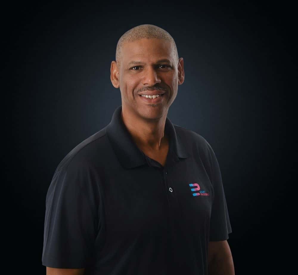 Chris Boudreaux - Live Event Production | Management | Event Producers - Live Event Production | Management | Event Producers Smiling man in a black polo shirt with a small colorful logo, posed against a dark studio backdrop.
