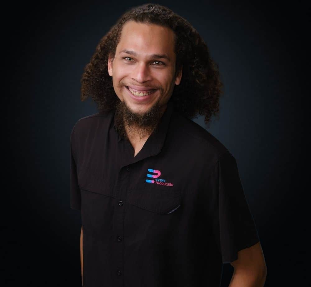 Daniel Brown - Live Event Production | Management | Event Producers - Live Event Production | Management | Event Producers Smiling man with long curly hair and a goatee in a black button-up shirt with an Event Producers logo against a dark background.
