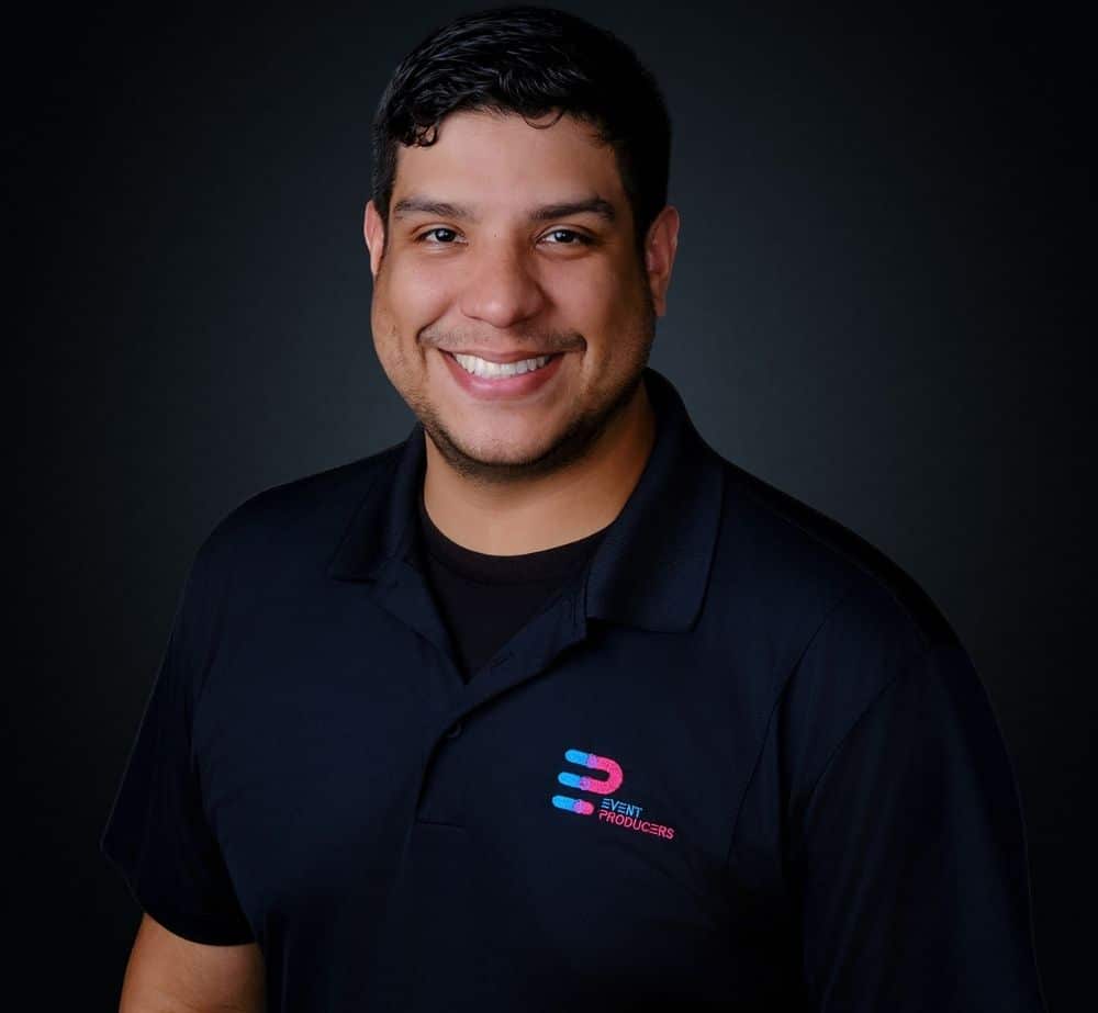 David Suazo - Live Event Production | Management | Event Producers - Live Event Production | Management | Event Producers Smiling man in a black polo shirt with the Event Producers logo on the left chest against a dark studio background.