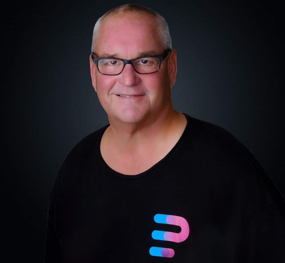 Eric Noble Sr - Live Event Production | Management | Event Producers - Live Event Production | Management | Event Producers Portrait of a smiling man with short gray hair and glasses, wearing a black shirt with a colorful logo on the chest, against a dark backdrop.