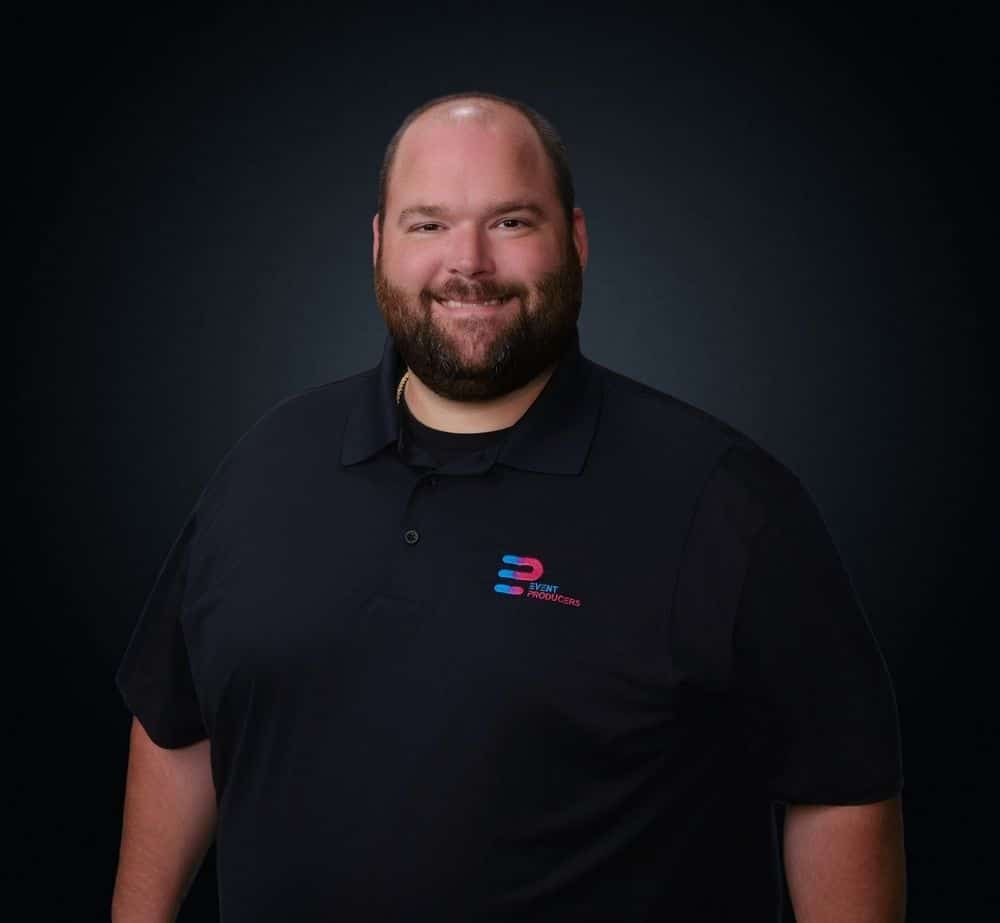 Eric Noble - Live Event Production | Management | Event Producers - Live Event Production | Management | Event Producers Smiling man with a beard in a black polo shirt with a blue‑red logo on the left chest, dark background.