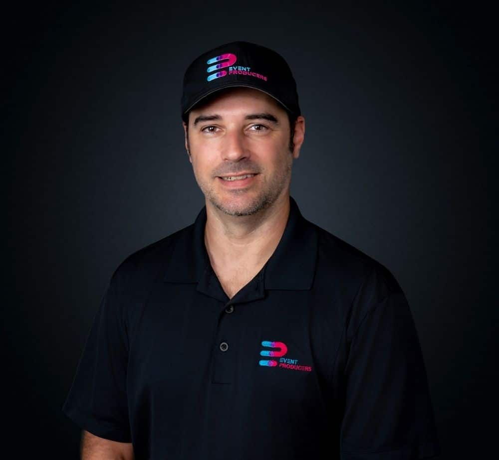 Justin Lucus - Live Event Production | Management | Event Producers - Live Event Production | Management | Event Producers Portrait of a man wearing a black polo shirt with an Event Producers logo on the chest and a matching cap against a dark gradient background.