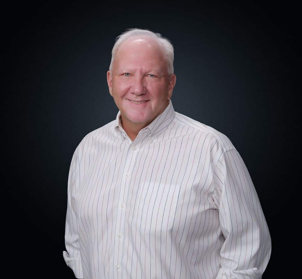 Keith Quick - Live Event Production | Management | Event Producers - Live Event Production | Management | Event Producers Portrait of a smiling older man with white hair wearing a white striped button-down shirt against a dark studio background