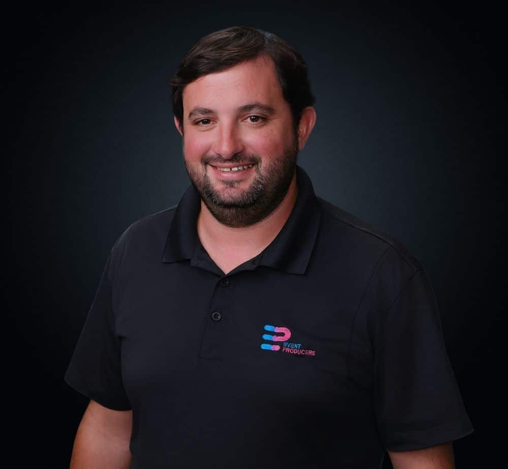 Logan Boudreaux - Live Event Production | Management | Event Producers - Live Event Production | Management | Event Producers Smiling man in a black polo shirt with a small colorful logo on the chest, photo portrait against a dark background