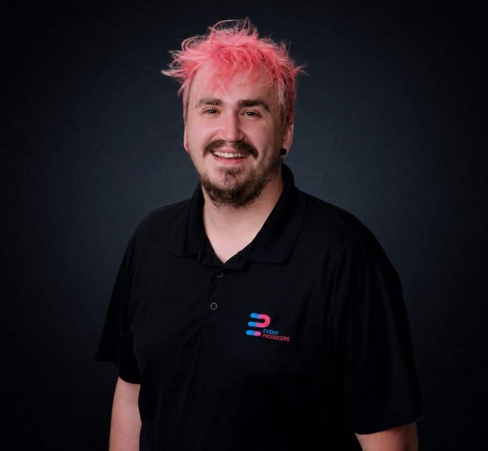 Nic McNulty - Live Event Production | Management | Event Producers - Live Event Production | Management | Event Producers Portrait of a smiling man with pink hair wearing a black polo shirt, logo on the left chest against a dark background.