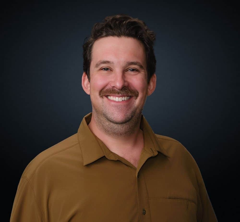 Ryan Nicholson - Live Event Production | Management | Event Producers - Live Event Production | Management | Event Producers Portrait of a smiling man with dark hair and a mustache, wearing a tan button-down shirt against a dark gradient backdrop.