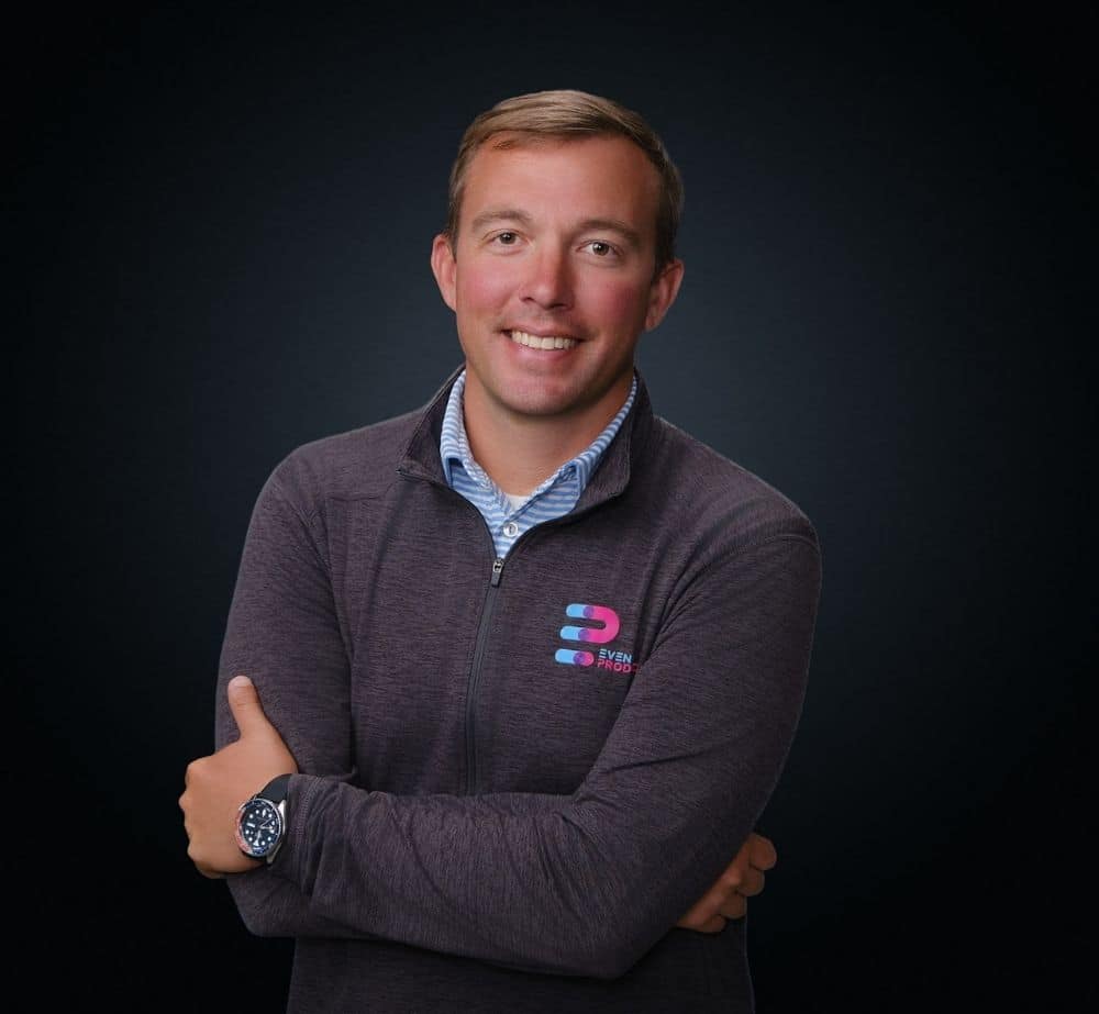 Ryan Quick - Live Event Production | Management | Event Producers - Live Event Production | Management | Event Producers Portrait of a smiling man with arms crossed, wearing a dark zip hoodie with a small logo on chest, against a dark studio background, watch visible.