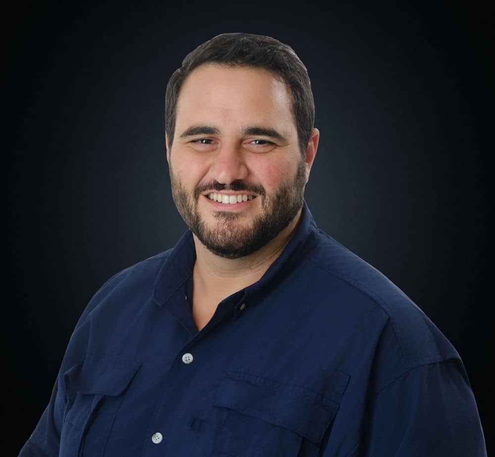 Untitled design 3 - Live Event Production | Management | Event Producers - Live Event Production | Management | Event Producers Head-and-shoulders portrait of a smiling man in a navy button-down shirt against a dark background.
