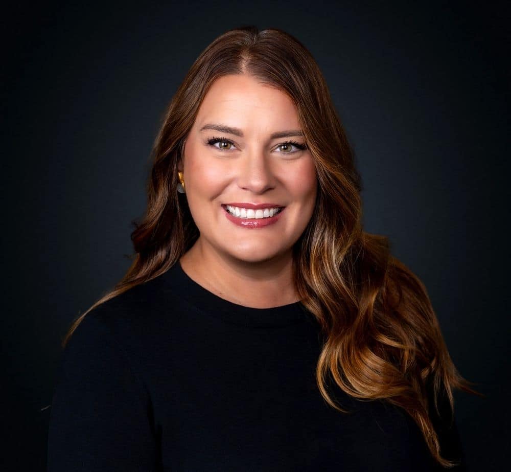 Whitney Hymel - Live Event Production | Management | Event Producers - Live Event Production | Management | Event Producers Head-and-shoulders portrait of a smiling woman with long brown hair in a black top against a dark backdrop.